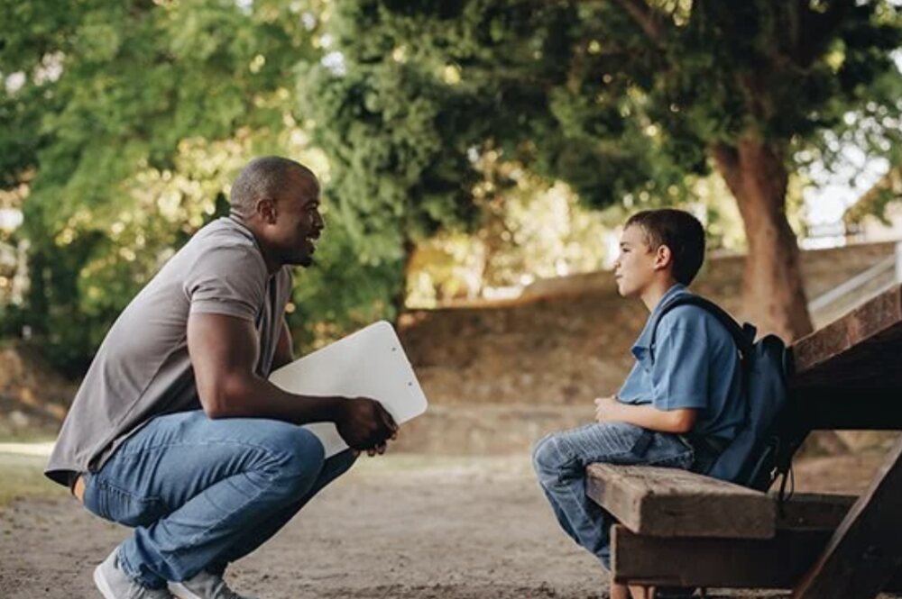 A mentor kneeling to speak with a young boy sitting on a park bench