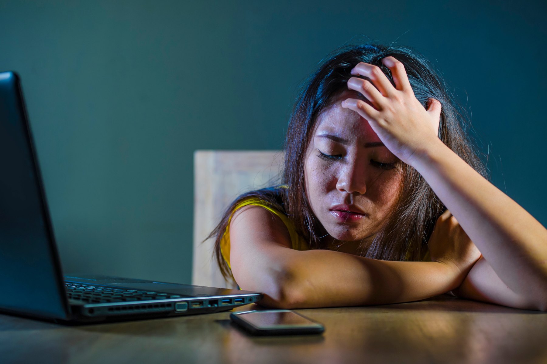 A woman resting her head on her arm at her laptop in a darkened room, emotionally drained