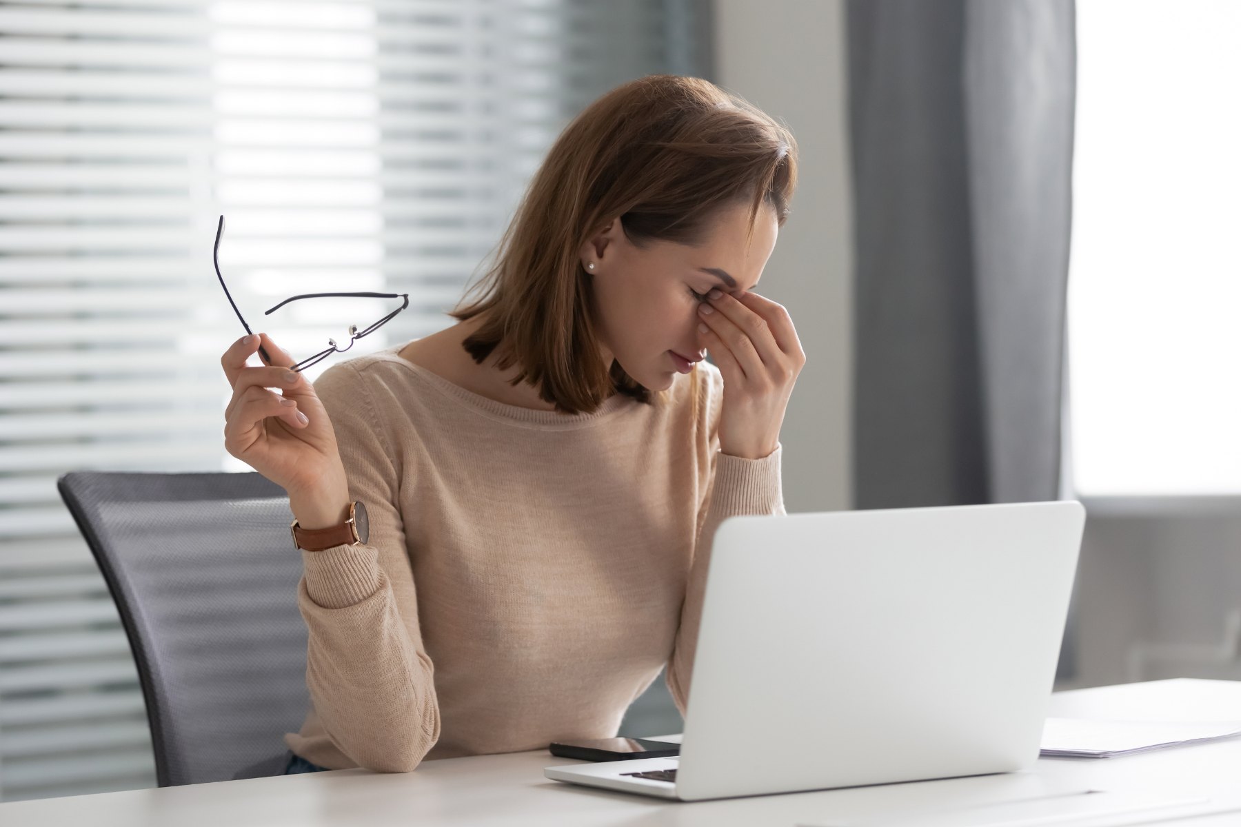 A woman removing her glasses and rubbing her eyes at her desk, showing screen fatigue