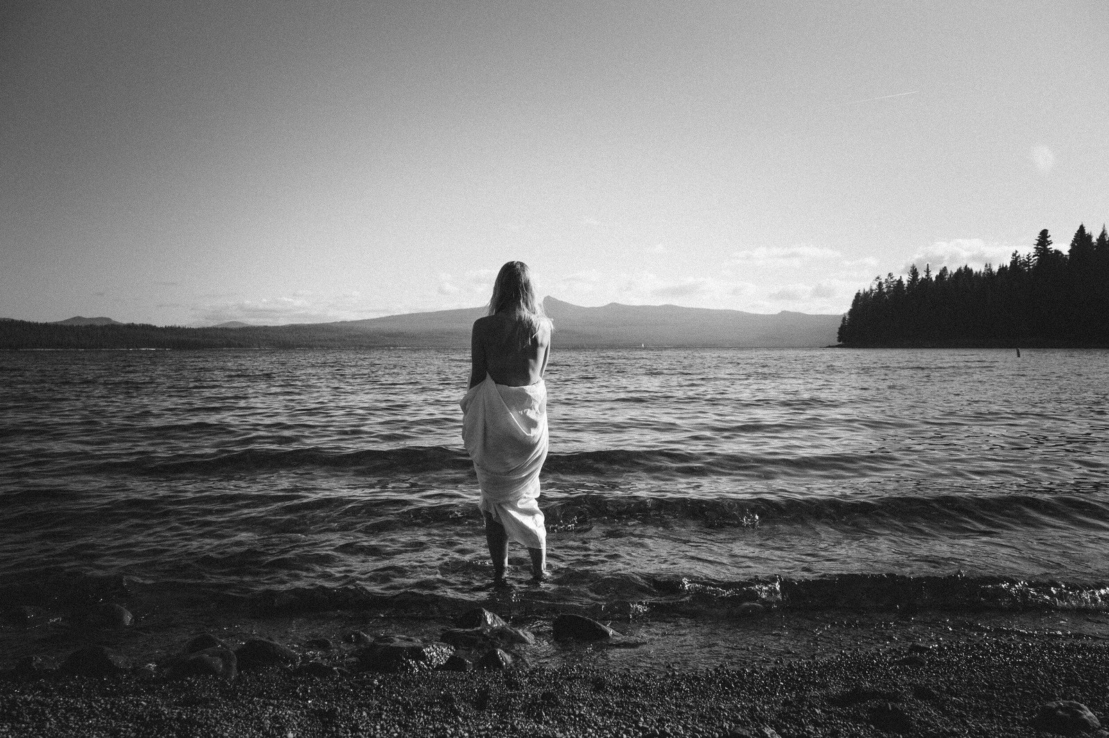 A woman standing at the edge of a still lake, facing the mountains