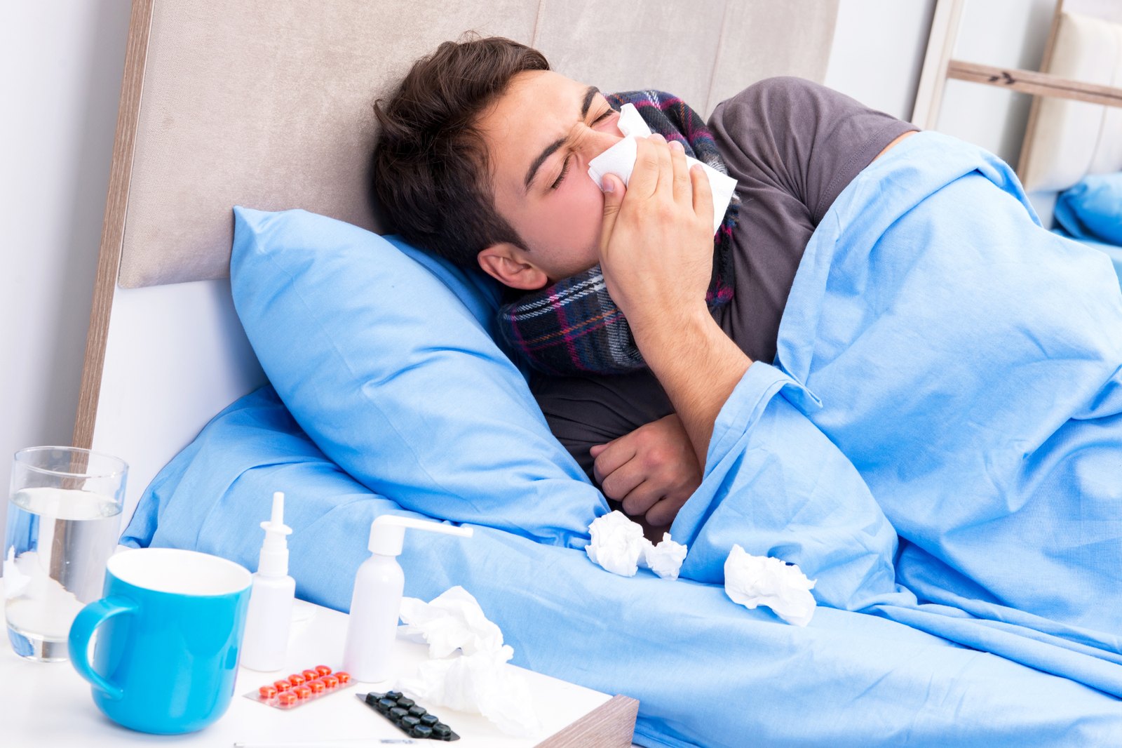 A man resting in bed surrounded by tissues, medicine, and a warm drink