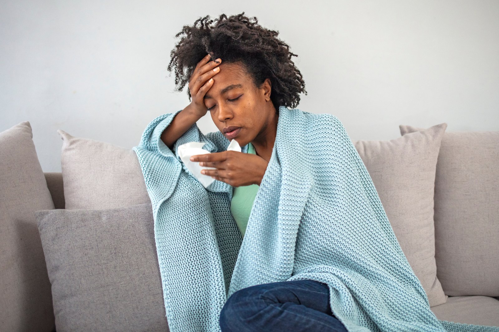 A woman wrapped in a teal blanket resting on the couch with a hand to her forehead